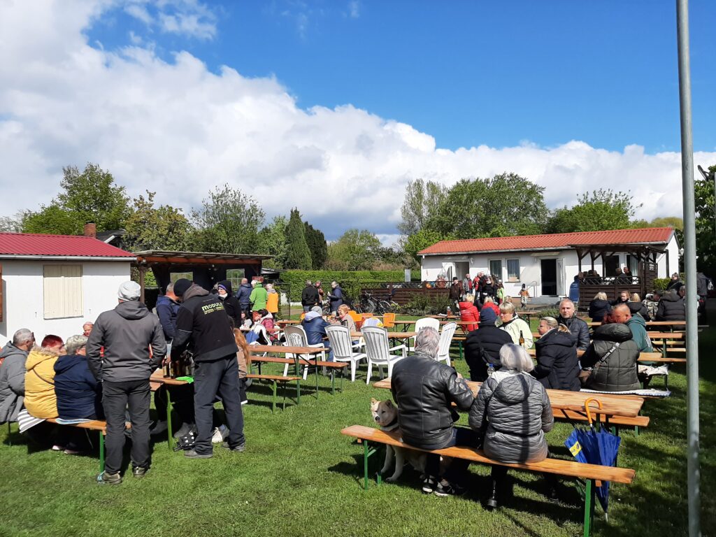Der Vereinsplatz des Kleingartenvereins Biesenhorst II. Links sieht man das Vereinsheim, rechts daneben den Grillunterstand und hinten rechts das Spartenheim. Auf dem Rasen sitzen Menschen an Biertischen, einige stehen am Grill an.