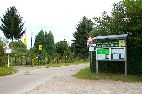 Der Eingang zur Kleingartenanlage Biesenhorst II. Man sieht einen Schaukasten mit diversen Aushängen mit dem Namensschild des Kleingartenvereins darüber. Im Hintergrund sieht man eine Parzelle und mehrere Verkehrsschilder.