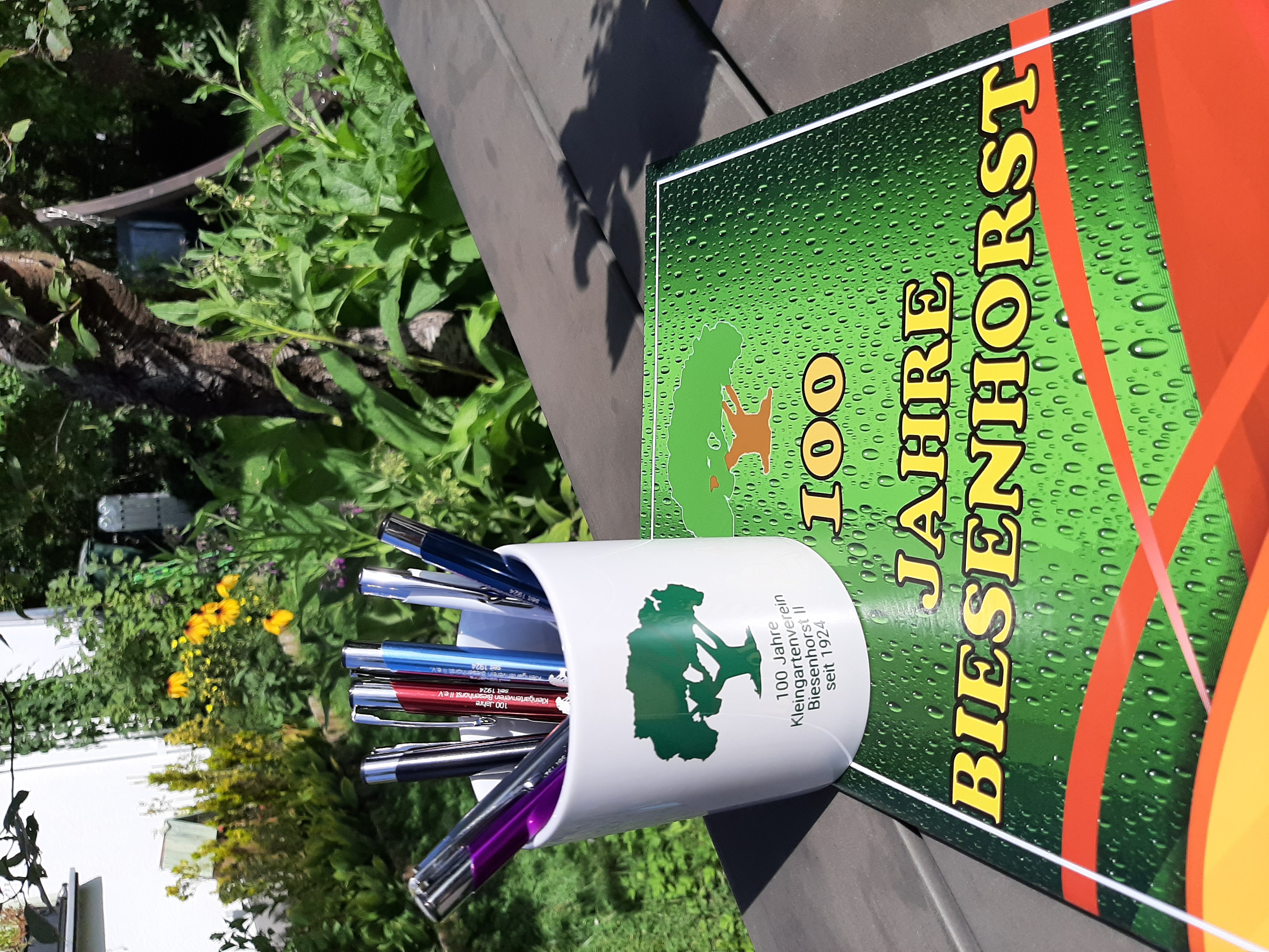 Foto der Festschrift zum 100jährigen Jubiläum der Kleingartenanlage Biesenhorst II. Diese liegt auf einem Tisch im Garten. Daneben steht die Jubiläums-Keramiktasse, in der mehrere bunte Jubiläums-Kugelschreiber stehen. Im Hintergrund sind Blumen zu sehen.